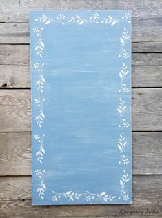 a blue painted canvas with white flowers and leaves on it, against a wooden background