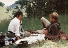 two people sitting on a blanket in the middle of a field with picnic food and drinks