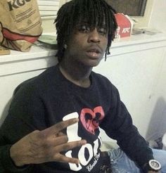 a man with dreadlocks sitting on the floor pointing to his left side while wearing a black shirt