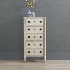 a white chest of drawers with flowers on top in front of a gray wall and wooden floor