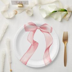 a white plate topped with a pink bow next to two gold forks and napkins