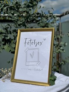 a sign that is sitting on top of a white table cloth and some plants in the background