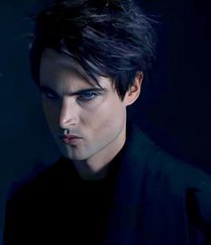 a man with black hair and blue eyes looks at the camera while standing in front of a dark background