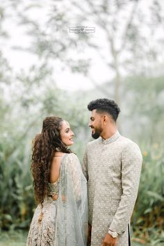 a man and woman standing next to each other in front of some bushes with trees