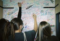 two girls writing on a wall covered in graffiti and stickers with their arms up