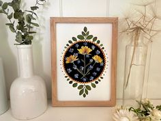 a white vase with some flowers in it next to a picture frame on a shelf