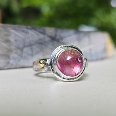 This tourmaline cabochon has a wonderful pink coloring with lots of interest and layers of depth. I set it in a bezel on a domed band with a high karat gold accent.  This smooth domed tourmaline is set in a fine silver bezel on a sterling silver backplate. The band is a half round domed shape that has been hammered to give it texture. A luxurious ball of solid 18K gold have been soldered to the band as an accent. The ring is marked with my makers mark, 925 and 18K. It is lightly oxidized to highlight the texture and the gold accents. It is a size 7 1/4. I was happy with how this ring evolved. It is a stunning ring with lots of panache! I hope you like it! Pink Round Cabochon Ruby Ring, Pink Cabochon Ruby Ring, Tourmaline Bezel Set Ring In Oval Cabochon, Tourmaline Ring With Bezel Setting In Oval Cabochon, Tourmaline Bezel-set Oval Cabochon Ring, Unique Pink Tourmaline Rings, Pink Tourmaline Sapphire Ring, Pink Round Tourmaline Jewelry, Pink Cabochon Ruby Ring Gift