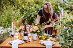 a woman sitting at a table with flowers and candles on it, surrounded by greenery