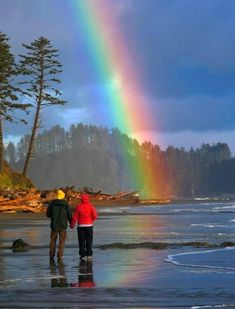 two people standing on the beach with a rainbow in the sky above them and trees behind them