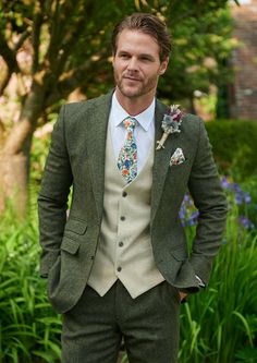 a man wearing a suit and tie standing in front of some bushes with his hands on his hips