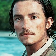 a man with long hair and a moustache looks at the camera while standing in front of water