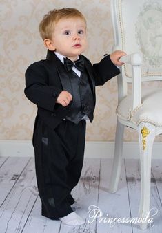 a little boy in a tuxedo standing next to a chair