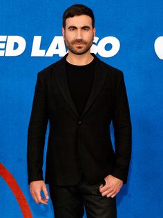 a man standing in front of a blue wall wearing a black suit and tie with his hands in his pockets