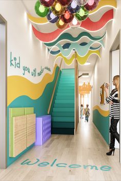 Statement wave effect ceiling and colourful ball lights with an Ombre effect staircase in turquoise. Kids you will make waves in a 3D font along the wall Kindergarten Design Interior, Daycare Entrance, School Wall Design, Kindergarten Entrance, School Wall Art Ideas, Childrens Ministry Decor