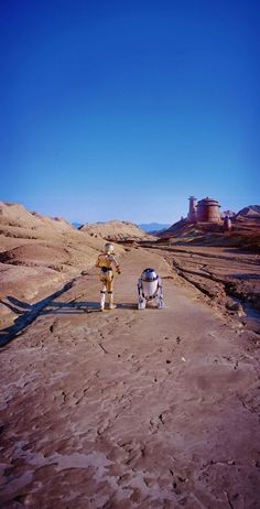 two people walking down a dirt road in the desert with a dog on their back