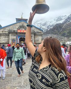 a woman is holding up a bell in front of a building with people walking around