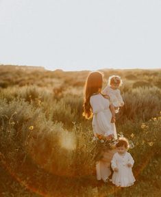 a woman holding two small children in a field with sun shining through the grass and flowers