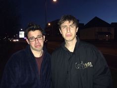 two men standing next to each other in front of a street at night with lights on