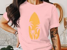 Embrace the summer vibes with this retro surfboard and palm trees graphic tee. Perfect for beach vacations, surfing adventures, or casual coastal living. This unique design brings a vintage beachy feel to your wardrobe. Ideal for both men and women who love seaside getaways and the sun-soaked surf culture. Available in short sleeve, long sleeve, and v-neck styles, this surf shirt is a must-have for your surf-inspired apparel collection.  Long Sleeve T-Shirt (Gilden 5400)  Fabrication: Fabric wei Tropical Printed Tops For Surfing, Tropical Printed Tops For Resortwear, Summer Surfing Screen Print T-shirt, Summer Surfing T-shirt With Letter Print, Beachy Summer Surfing T-shirt, Summer Beachy T-shirt For Surfing, Beachwear Pink Tops With Graphic Print, Pink Graphic Print Beachwear Tops, Pink Beachwear Tops With Graphic Print