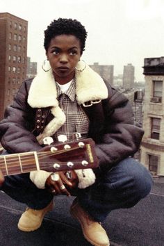 a man sitting on top of a roof with an acoustic guitar in his hand and buildings in the background