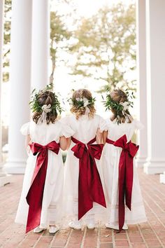 three girls in white dresses with red sashes and flower crowns on their heads are looking at each other