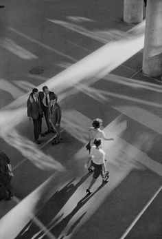 black and white photograph of people walking down the street in front of columns with shadows on them