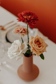 three flowers in a vase sitting on a table with plates and wine glasses behind them