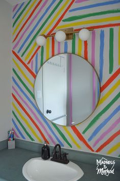 a bathroom sink under a round mirror in front of a colorful wall