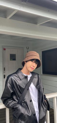 a young man wearing a leather jacket and hat, standing on a porch with his hands in his pockets