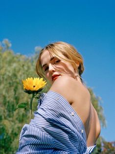 a woman in a blue dress holding a yellow flower and looking at the camera while standing outside