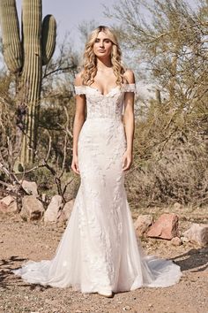 a woman standing in front of a cactus wearing a wedding dress with off the shoulder sleeves