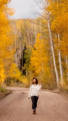 a woman walking down a dirt road in front of trees with yellow leaves on them