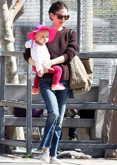 a woman holding a baby in her arms while walking down a sidewalk next to a fence