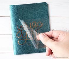 a hand holding a pen next to a green book with gold lettering on the cover