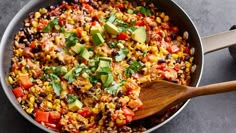 a skillet filled with corn, avocado and other vegetables next to a wooden spoon