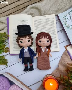 two knitted dolls sitting next to an open book on a white wooden table with candles