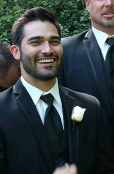 two men in suits and one is wearing a rose boutonniere, smiling at the camera