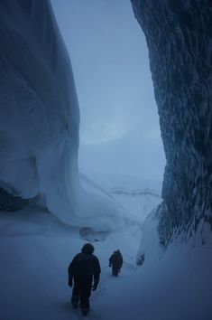 two people walking up a snow covered mountain in front of an ice cave on a cloudy day