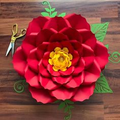 a red paper flower sitting on top of a wooden floor next to scissors and green leaves