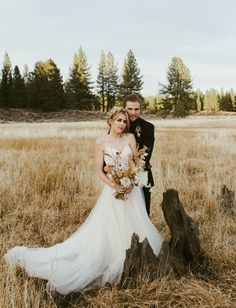 a bride and groom standing in the middle of a field with tall grass, surrounded by trees