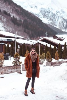 a woman standing in the snow wearing a brown fur coat and black top with her hands on her hips