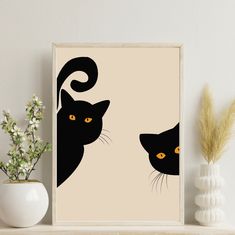 two black cats with yellow eyes on a beige background framed in a wooden frame next to a plant