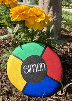 a rock with the word simon painted on it next to yellow flowers and dirt in front of a tree