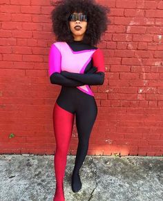 a woman in pink and black bodysuit standing next to a brick wall with her arms crossed