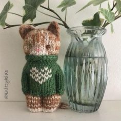 a knitted teddy bear sitting next to a vase with leaves in it and a green sweater on