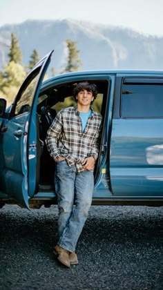 a man standing in the open door of a blue pickup truck with mountains in the background