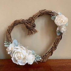 a heart shaped wreath with white flowers and leaves hanging on a wall next to a wooden table