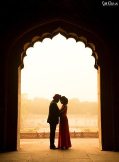 a man and woman standing in front of an arch with the sun setting behind them