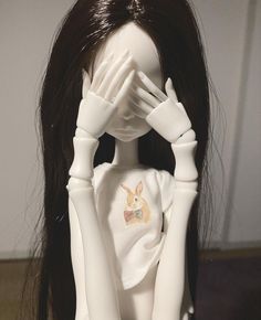 a white doll with long black hair covering her eyes and hands behind her head, in front of a wall