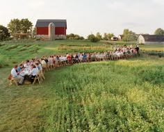 a long line of people sitting at tables in the middle of a field with farm buildings behind them